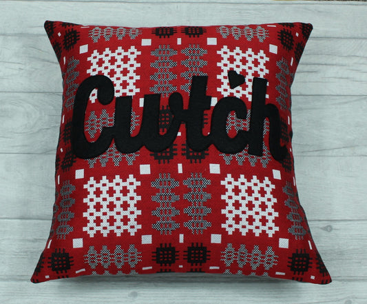 A square-shaped cushion with a red and gray Welsh tapestry pattern and the word 'CWTCH' appliqued in black.