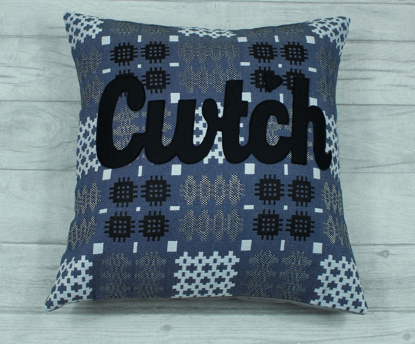 A blue coloured square-shaped cushion in a tradional  Welsh tapestry pattern fabric with the word  'CWTCH' appliqued in black hand cut felt