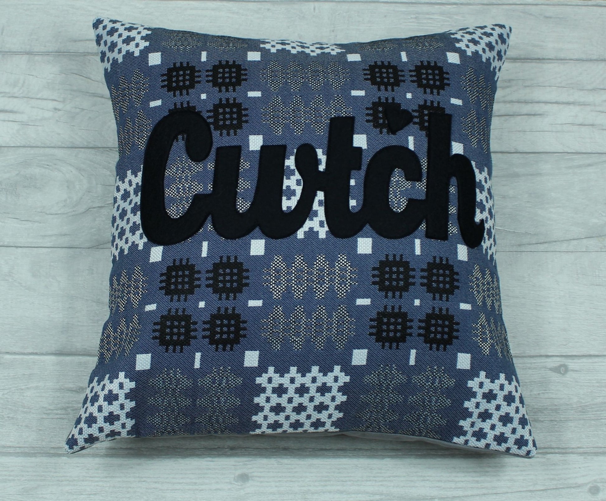 A blue coloured square-shaped cushion in a tradional  Welsh tapestry pattern fabric with the word  'CWTCH' appliqued in black hand cut felt