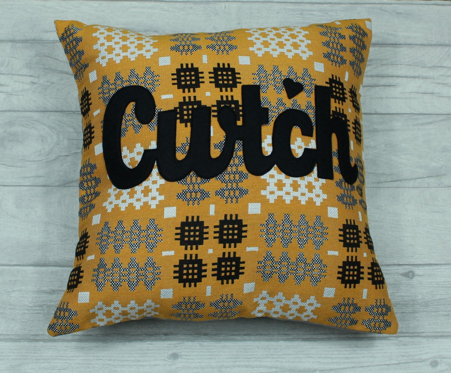 A gold coloured square-shaped cushion  in a tradional  Welsh tapestry pattern fabric with the word  'CWTCH' appliqued in black hand cut felt