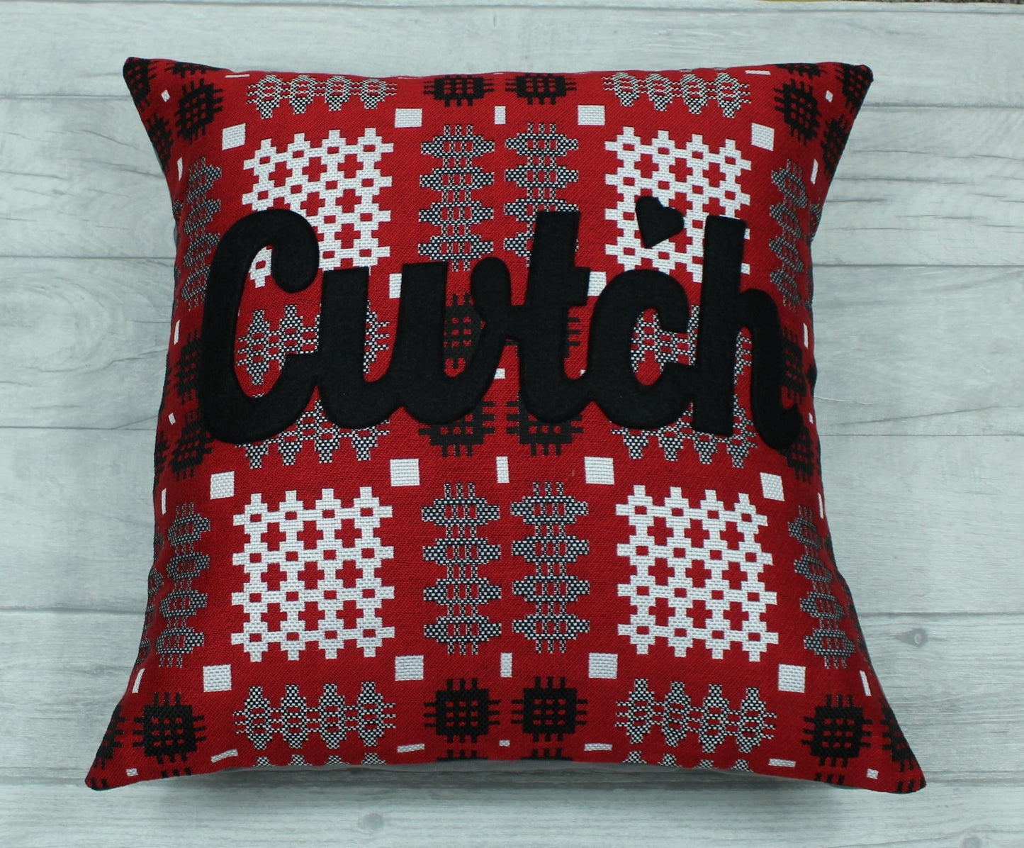 A red coloured square-shaped cushion in a tradional  Welsh tapestry pattern fabric with the word  'CWTCH' appliqued in black hand cut felt