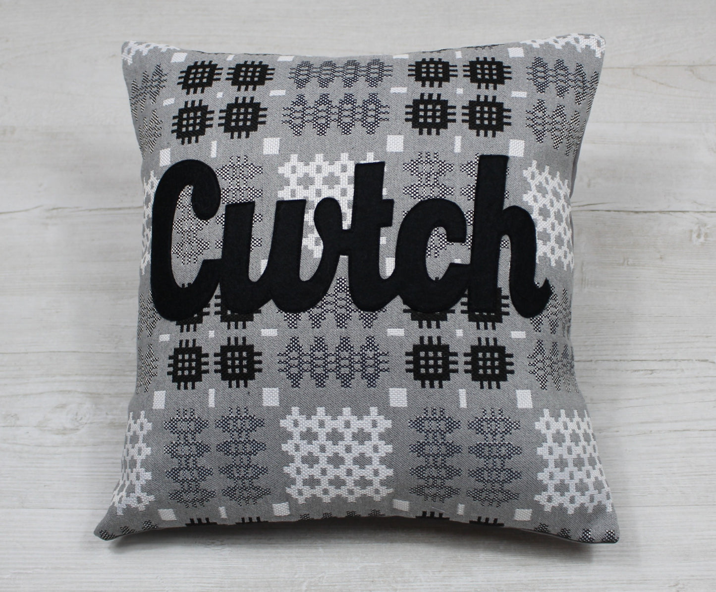 A silver coloured square-shaped cushion in a tradional  Welsh tapestry pattern fabric with the word  'CWTCH' appliqued in black hand cut felt